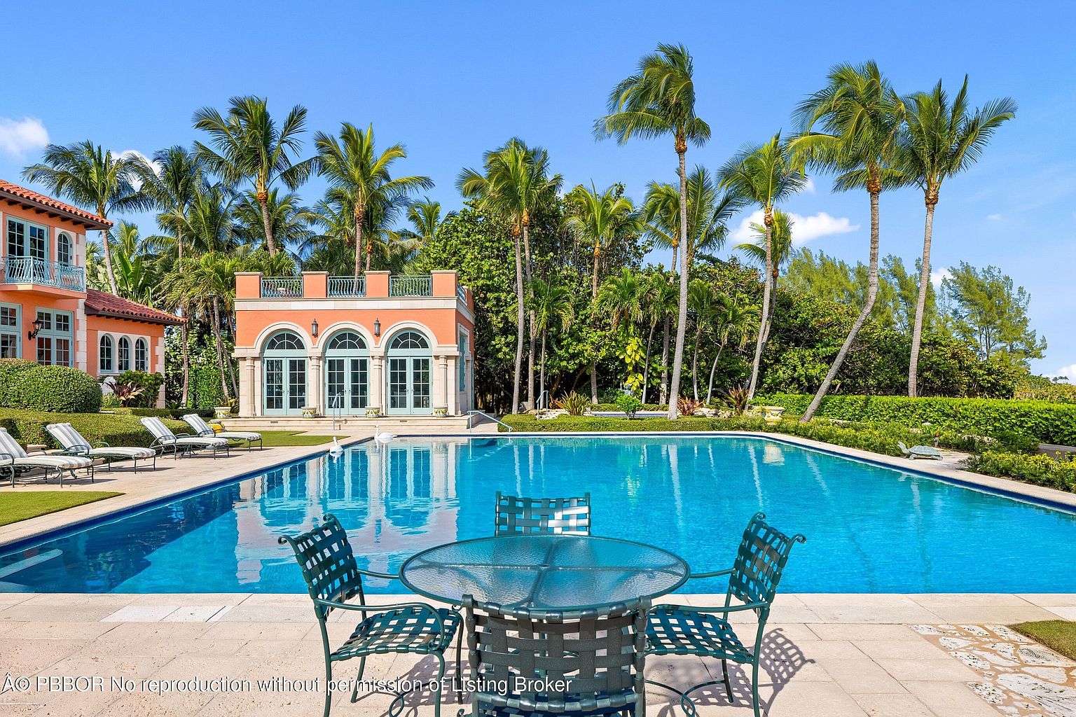 This image showcases a luxurious outdoor pool area, featuring a large, inviting pool with clear blue water. A patio set with a glass table and metal chairs sits in the foreground, while a charming pool house with arched windows and a balcony stands in the background. Palm trees surround the area, creating a tropical and serene atmosphere, perfect for relaxation and entertainment.