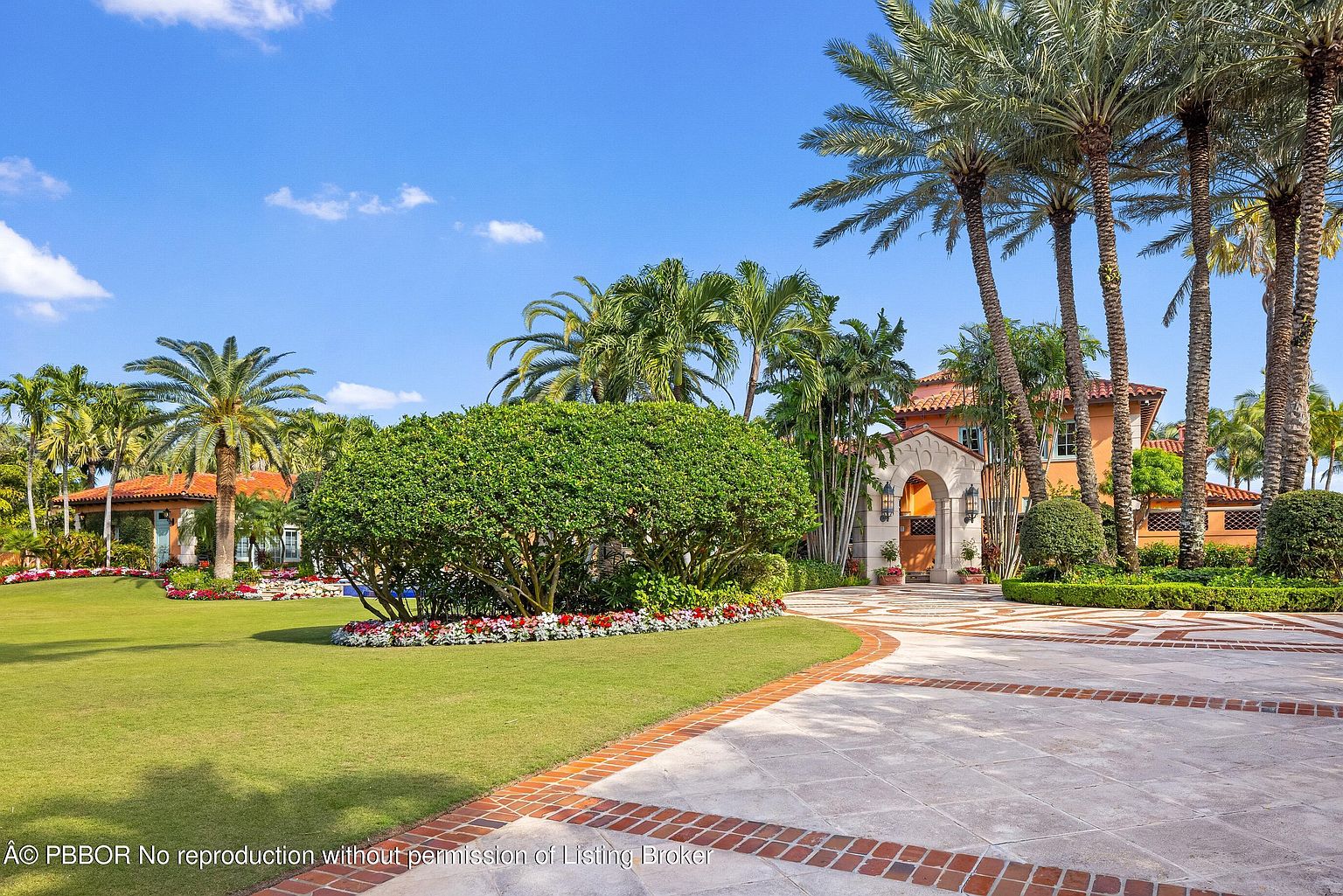This image showcases a beautifully landscaped yard with a grand driveway leading to a luxurious home. The lush green lawn is complemented by vibrant flower beds and mature palm trees, creating a tropical paradise. The driveway is made of stone with brick accents, adding to the property's curb appeal and overall elegance.