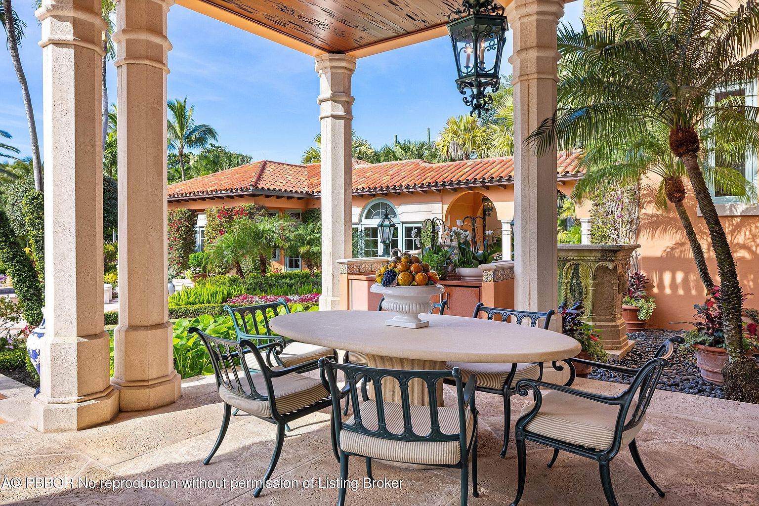 This image showcases an elegant outdoor patio area, featuring a stone dining table with chairs under a covered structure supported by columns. The patio is adorned with lush greenery and landscaping, with a view of a Mediterranean-style house in the background. The scene exudes a sense of luxury and relaxation, perfect for outdoor entertaining.