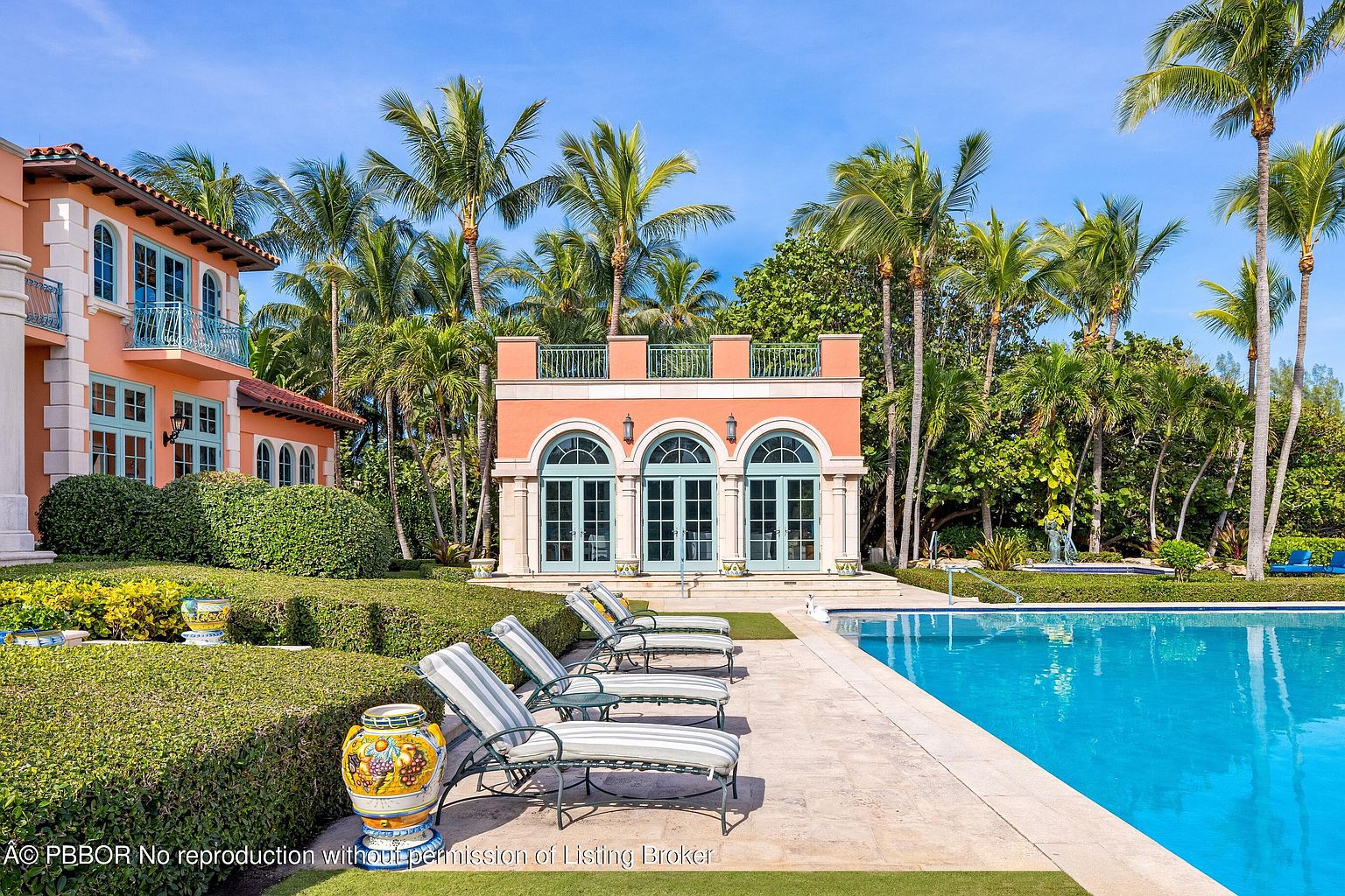 This image showcases a luxurious outdoor pool area, featuring a pristine blue pool bordered by a stone patio and several lounge chairs. A charming pool house with arched doorways and a balcony stands in the background, surrounded by lush tropical landscaping including palm trees and manicured hedges. The overall impression is one of relaxation and upscale living.