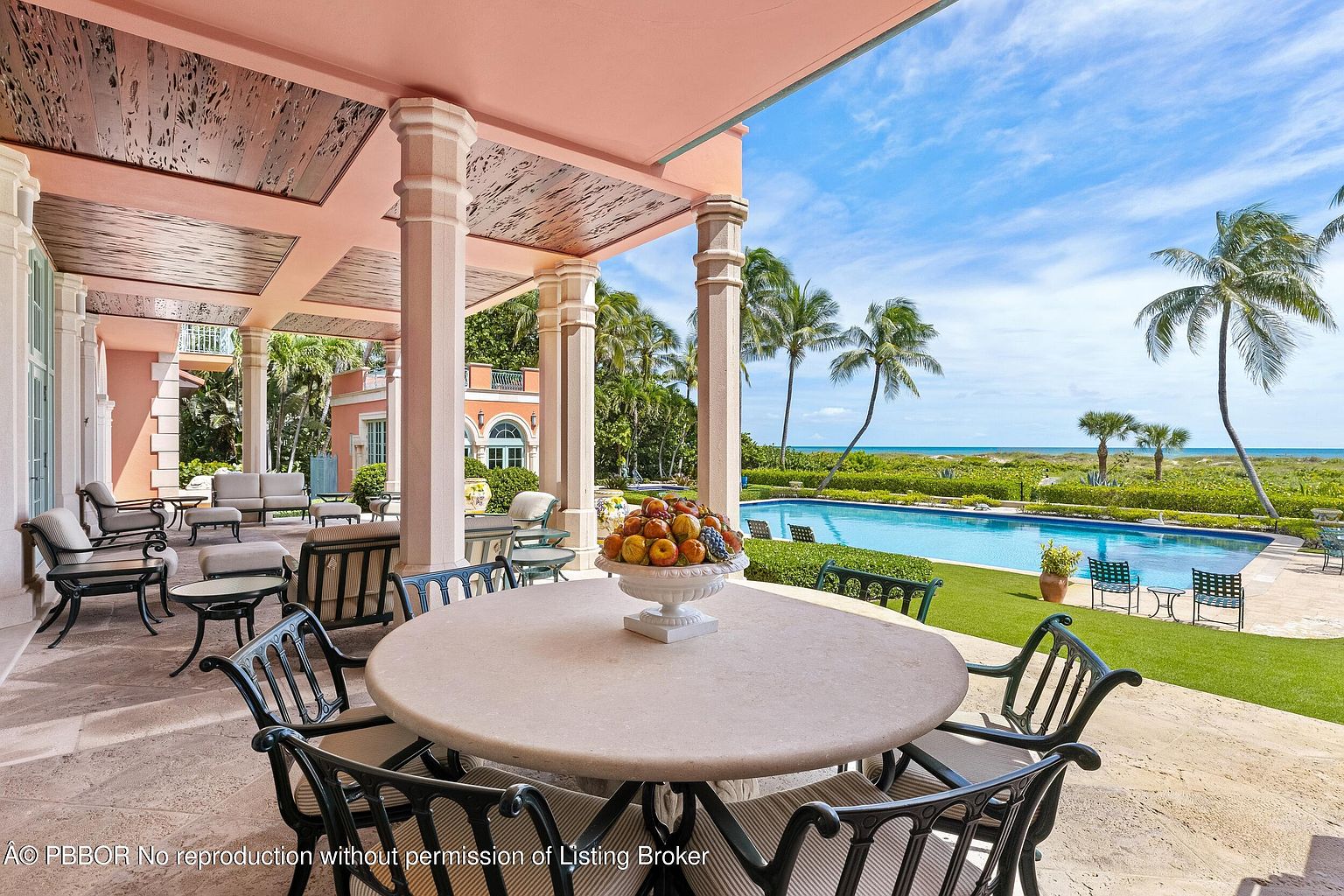This image showcases a luxurious outdoor patio area with a large round table and chairs, perfect for dining and entertaining. The patio is covered by a stylishly designed roof supported by columns, offering shade and architectural interest. Beyond the patio, a well-maintained lawn leads to a swimming pool and a view of the ocean, framed by palm trees, creating a serene and upscale ambiance.