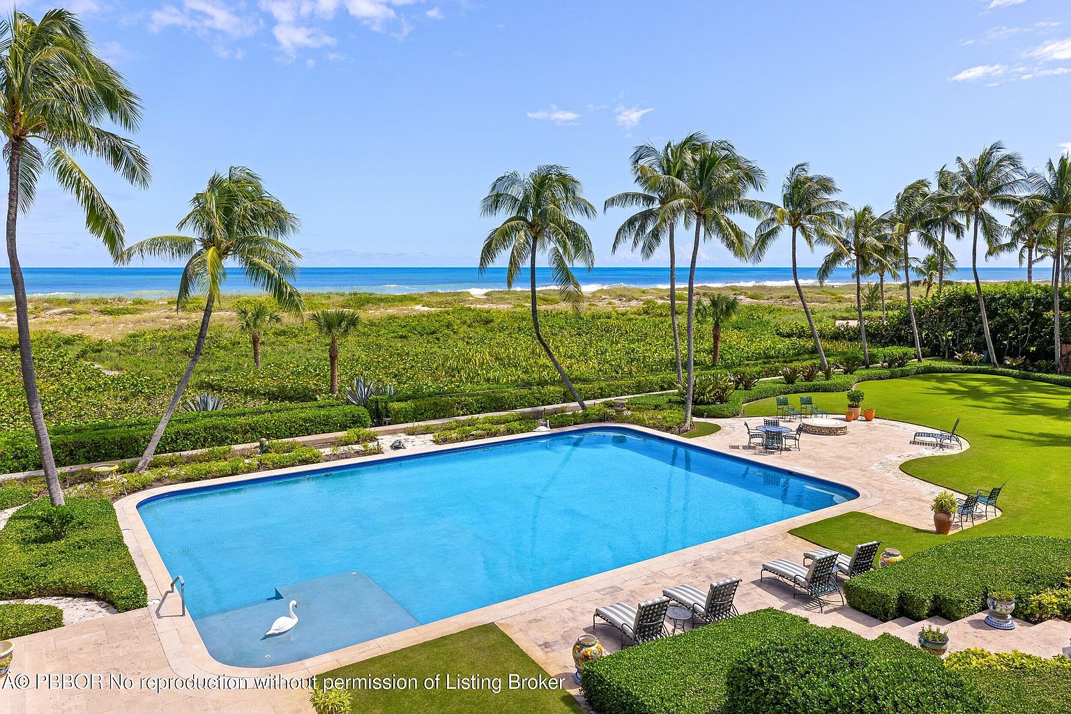 This image showcases a luxurious backyard featuring a large rectangular swimming pool with a swan float. The pool is surrounded by a stone patio, lounge chairs, and meticulously manicured greenery, including palm trees and hedges. Beyond the yard, there is a view of the beach and ocean, creating a serene and upscale atmosphere.