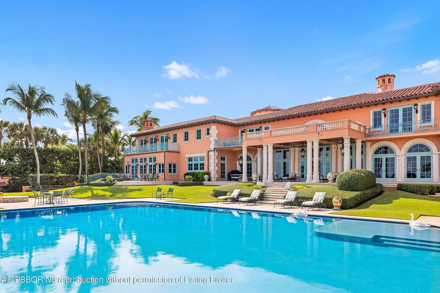 This image showcases a luxurious backyard featuring a large swimming pool with crystal clear water reflecting the sky. The pool is surrounded by well-manicured green lawns, lounge chairs, and elegant landscaping. A grand, multi-story Mediterranean-style house with peach-colored walls and blue-green window frames stands in the background, adding to the overall impression of opulence and relaxation.