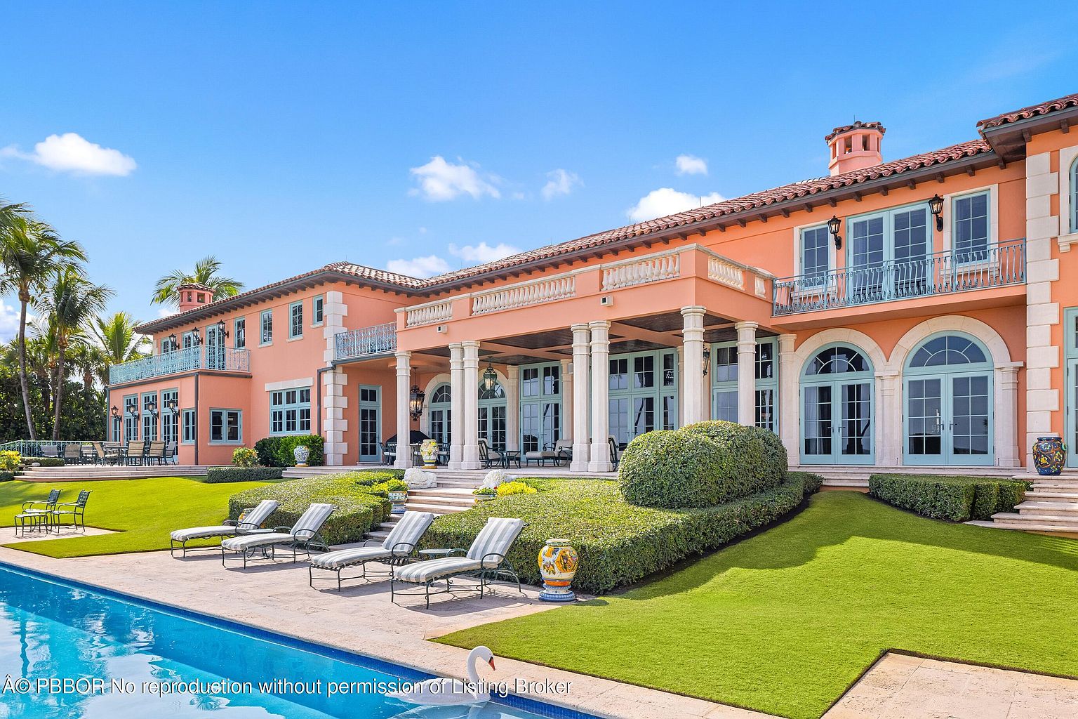 This image showcases the rear exterior of a luxurious Mediterranean-style home, emphasizing its stunning pool area. The pool is surrounded by lounge chairs and manicured landscaping, with the grand facade of the house featuring arched windows, columns, and balconies. The overall impression is one of opulence and relaxation, perfect for upscale living.