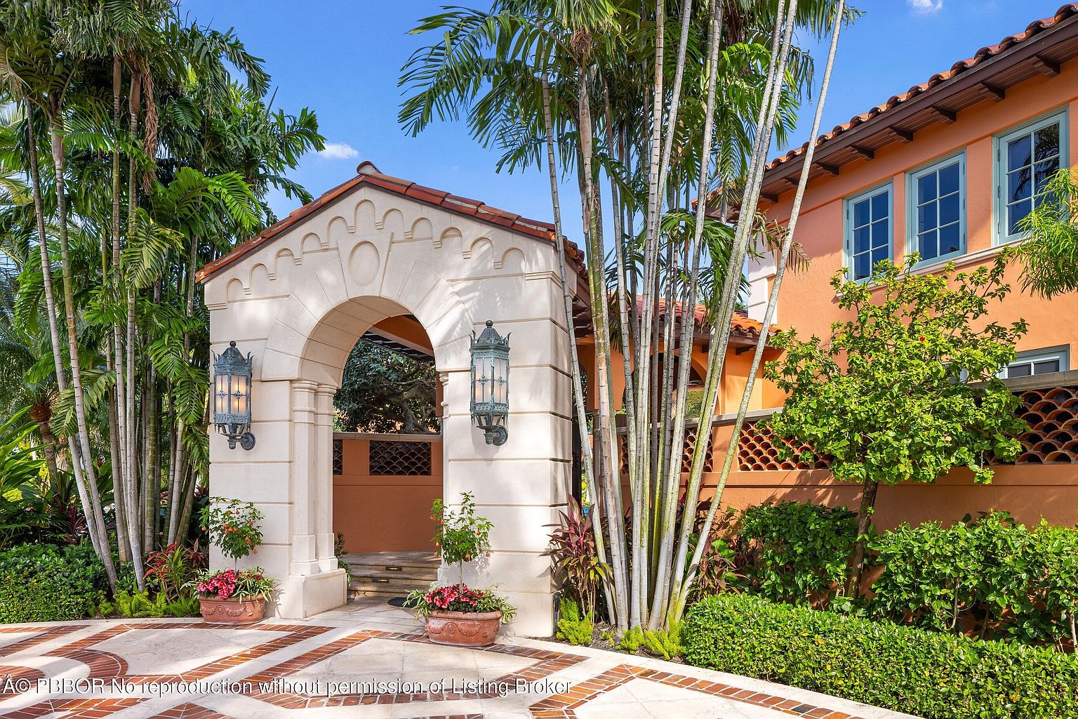 This image showcases a grand entryway to a Mediterranean-style home. The entrance features a stone archway with decorative lighting fixtures, leading to a courtyard or garden area. Lush greenery and palm trees surround the entrance, adding to the property's curb appeal and creating a sense of privacy and luxury.