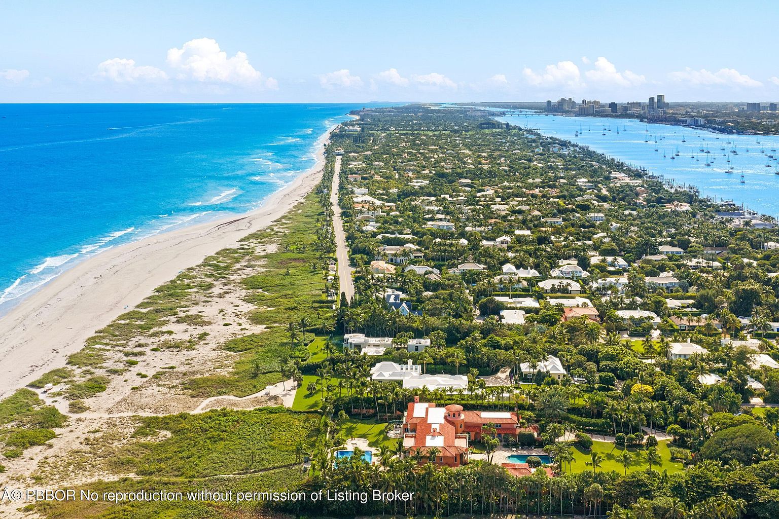 This aerial shot showcases a luxurious coastal property with a large, red-roofed mansion as its focal point, complete with a pool and manicured grounds. The property is situated on a narrow strip of land between the ocean and a bay, surrounded by lush greenery and other upscale homes. The image emphasizes the property's prime location and exclusive setting.