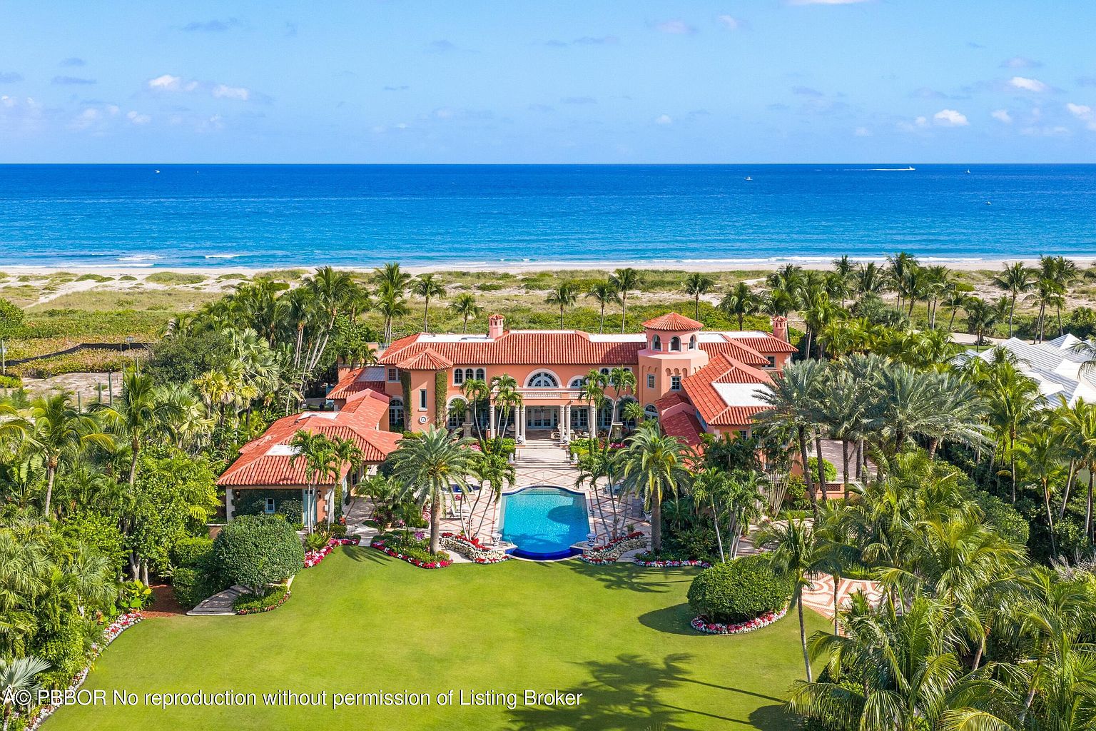 This aerial view showcases a luxurious beachfront estate with a terracotta-tiled roof, a central courtyard, and a large swimming pool. Lush landscaping with palm trees surrounds the property, leading to a pristine beach and the ocean. The overall impression is one of opulence and privacy.