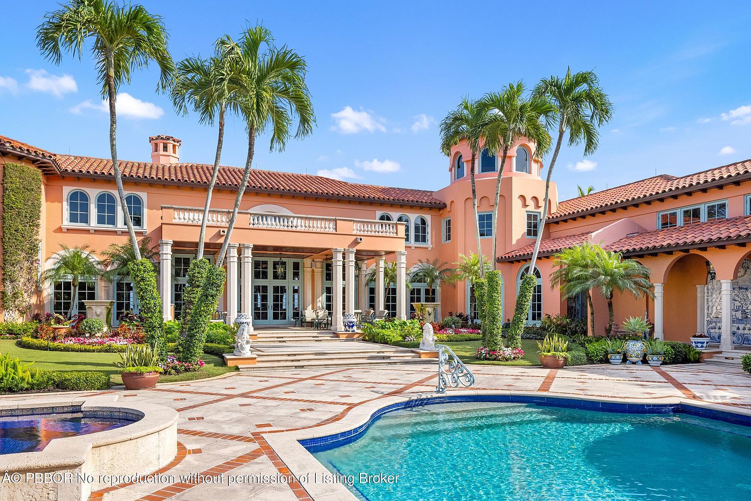 This image showcases a luxurious Mediterranean-style estate with a focus on the outdoor pool and patio area. The architecture features a terracotta roof, peach-colored stucco walls, and elegant columns supporting a balcony. Lush landscaping, including palm trees and manicured gardens, surrounds the property, creating a serene and upscale ambiance. The pool's clear blue water invites relaxation and leisure.