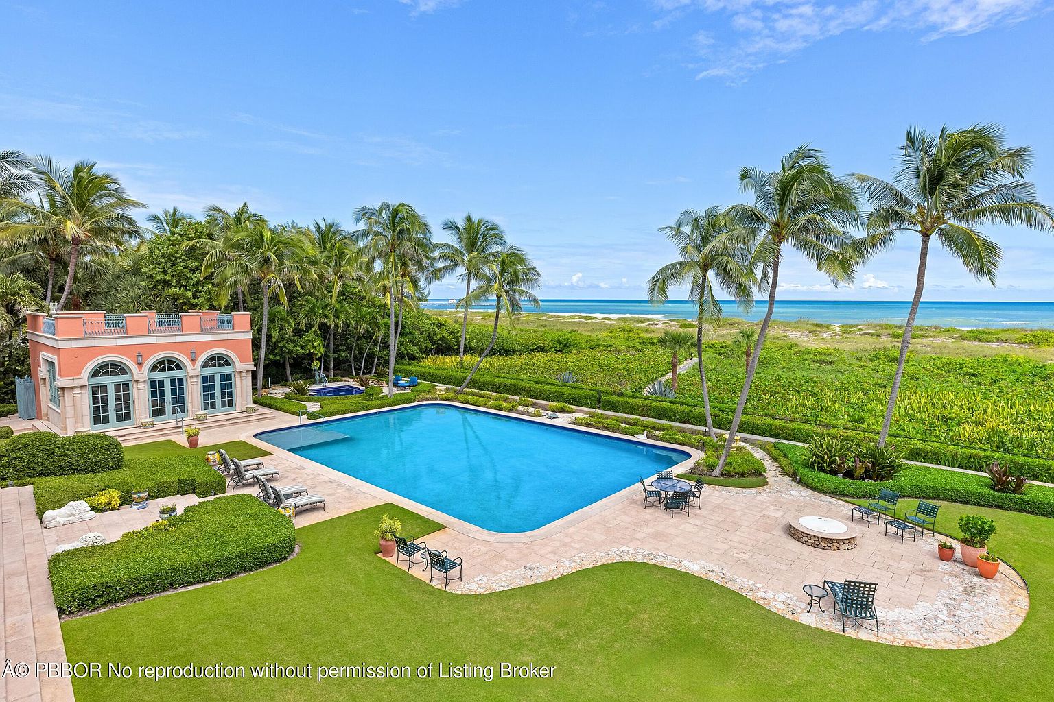 This is an aerial view of a luxurious backyard featuring a large rectangular swimming pool, a separate spa, and a charming pool house with arched windows. The property boasts meticulously manicured lawns, lush tropical landscaping with palm trees, and a clear view of the ocean beyond the dunes. The scene evokes a sense of tranquility and upscale coastal living.