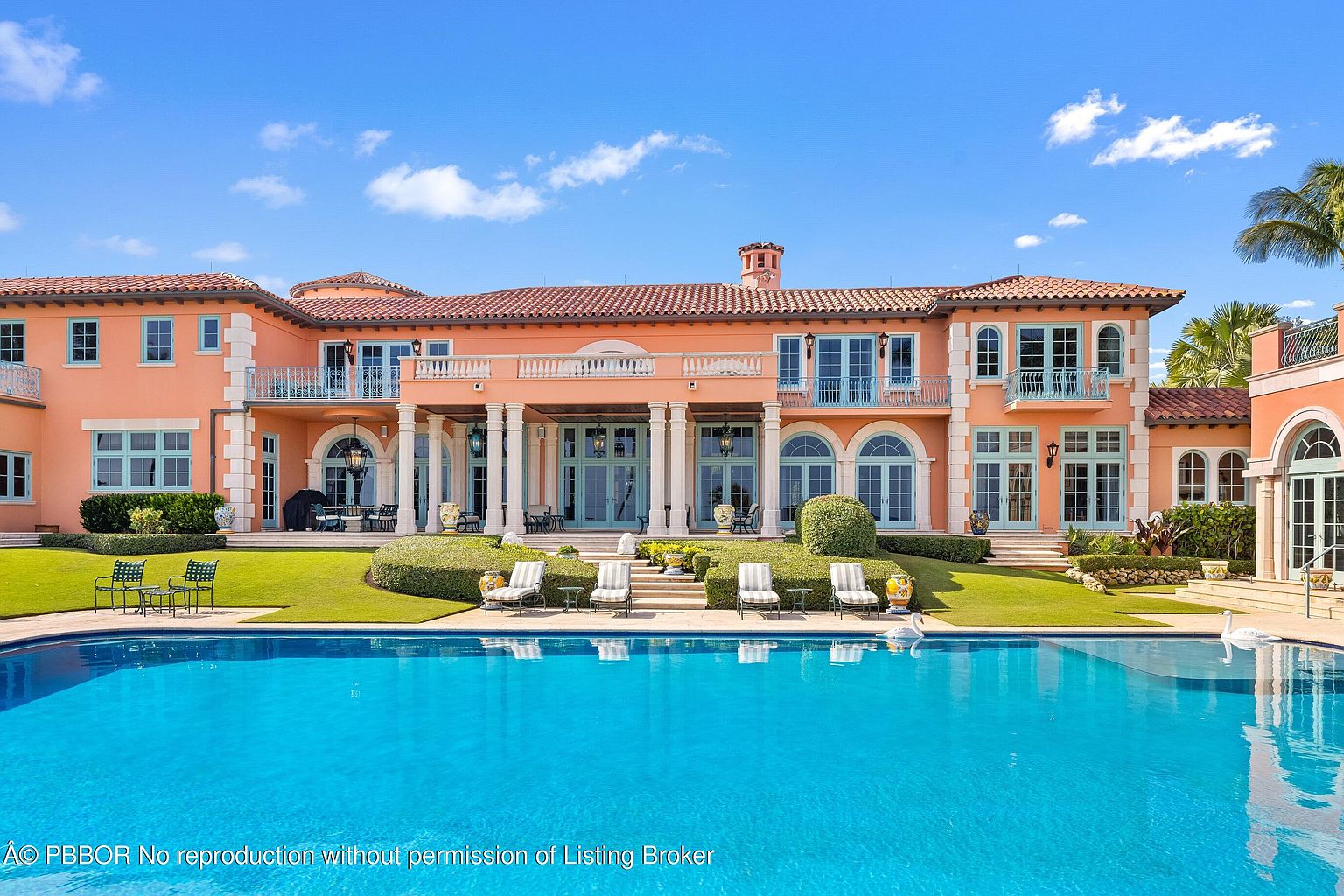 This image showcases the rear exterior of a luxurious Mediterranean-style home, emphasizing its stunning pool area. The expansive pool, with its clear blue water, is the focal point, reflecting the elegant architecture of the house. The property features manicured lawns, decorative shrubbery, and outdoor lounge furniture, creating an inviting and upscale outdoor living space.