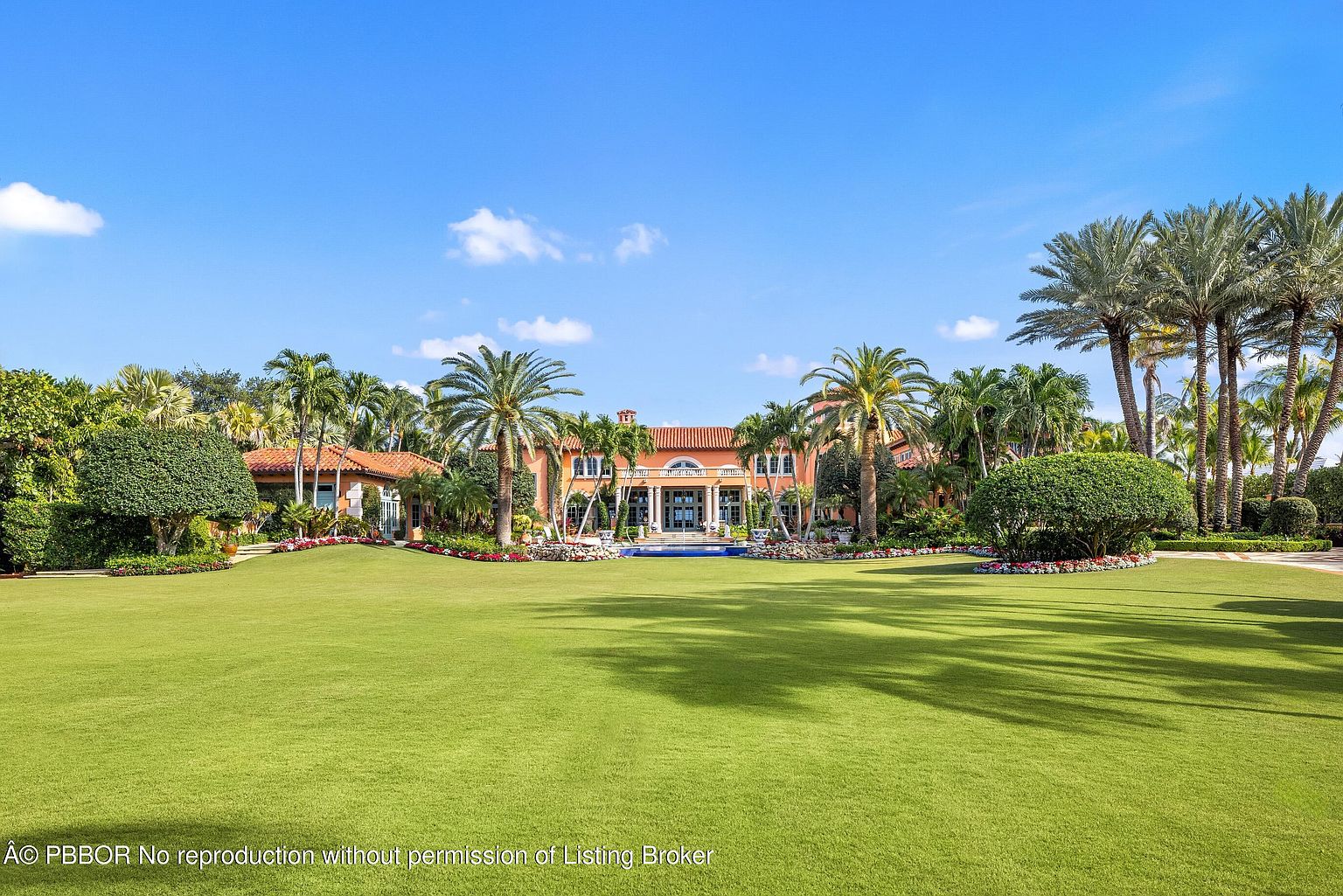 This is a front exterior view of a grand estate, showcasing a large manicured lawn leading up to a luxurious Mediterranean-style home. The property features lush landscaping with various palm trees and manicured shrubs, adding to the overall curb appeal. The house itself has a symmetrical design with a terracotta roof, columns, and a central entrance, suggesting high-end living and architectural elegance.