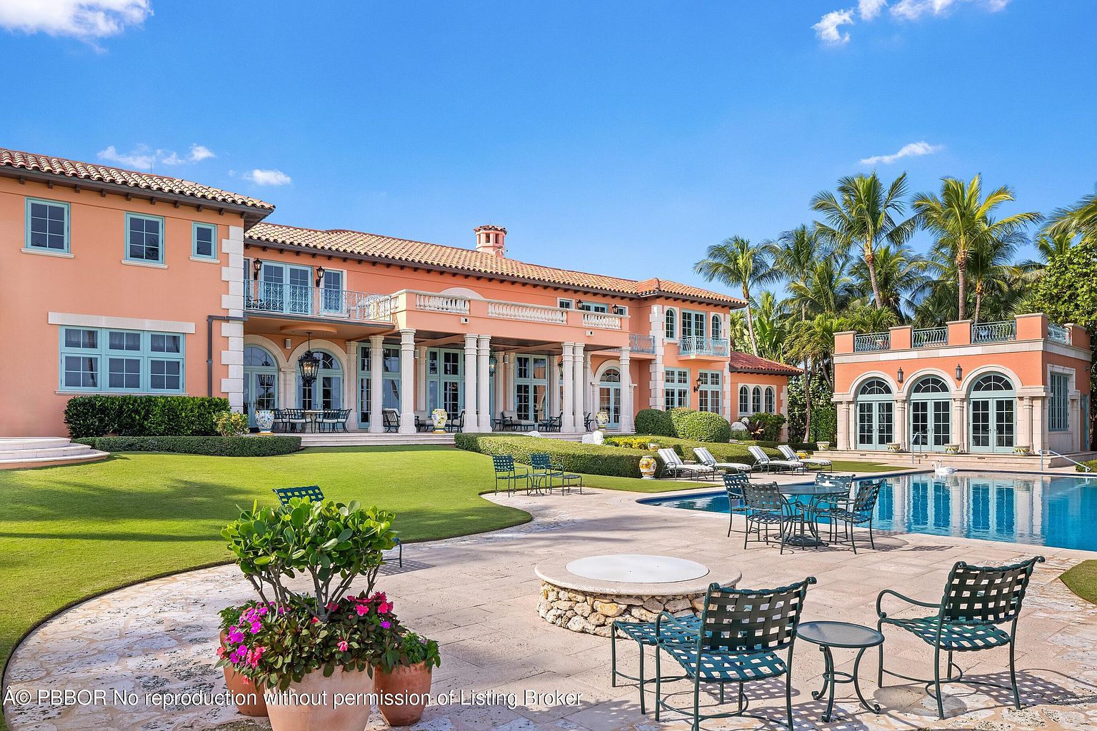 This image showcases the rear exterior of a luxurious Mediterranean-style home, emphasizing the outdoor living space. A pristine pool is surrounded by patio furniture and a fire pit, with lush green lawns and manicured landscaping extending towards the house. The architecture features coral-colored walls, white columns, and multiple balconies, creating an inviting and elegant atmosphere.