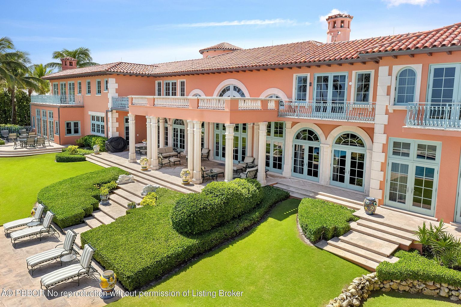 This is a rear view of a luxurious, multi-story Mediterranean-style home. The exterior features a salmon-colored facade, a red tile roof, and multiple balconies with light blue railings. The manicured lawn includes tiered landscaping with neatly trimmed hedges and stone steps leading up to the house, creating an inviting outdoor space.