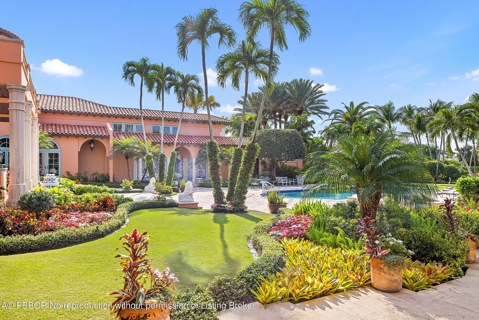 This exterior shot showcases a meticulously landscaped yard and garden, featuring lush green lawns, vibrant flowerbeds, and mature palm trees. The property includes a swimming pool in the background, suggesting a luxurious and private outdoor space. The overall impression is one of tranquility and sophisticated outdoor living.
