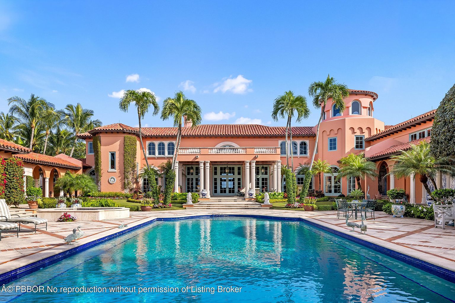 This image showcases a luxurious estate with a large swimming pool in the foreground. The pool's clear blue water reflects the grand facade of the house, which features a terracotta roof, multiple columns, and arched windows. Palm trees and manicured landscaping surround the pool area, creating a serene and upscale atmosphere.