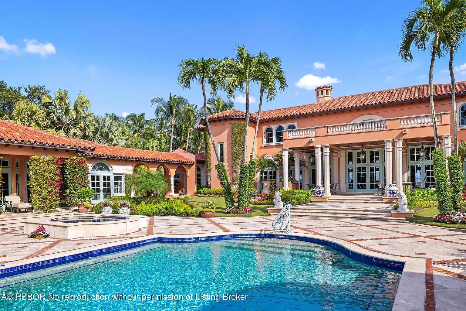 This image showcases the rear exterior of a luxurious Mediterranean-style home, emphasizing the pool and surrounding landscape. The pool features a blue tile interior and is bordered by a stone patio with brick accents. The house boasts a terracotta tile roof, peach-colored stucco walls, and lush landscaping with palm trees, creating a resort-like atmosphere perfect for relaxation and entertainment.