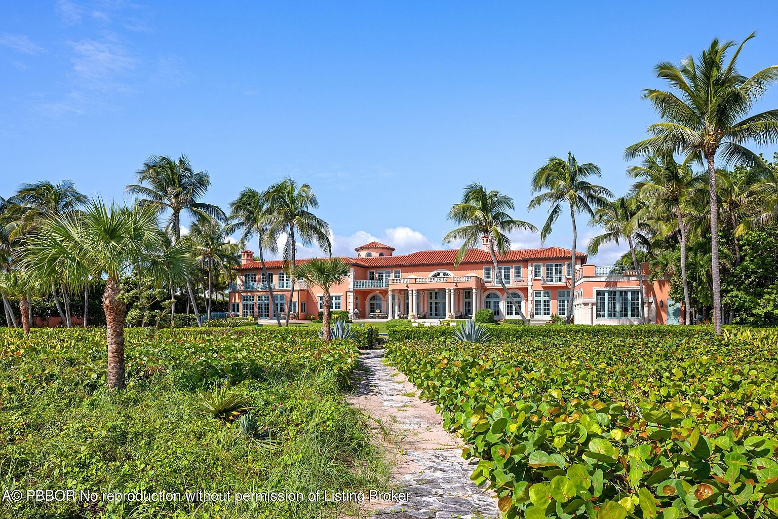 This image showcases the grand front exterior of a luxurious estate. The building features a terracotta roof, a symmetrical facade with multiple windows, and a colonnaded entrance. Lush landscaping, including palm trees and manicured greenery, surrounds the property, leading to a stone pathway that invites viewers to explore the residence.