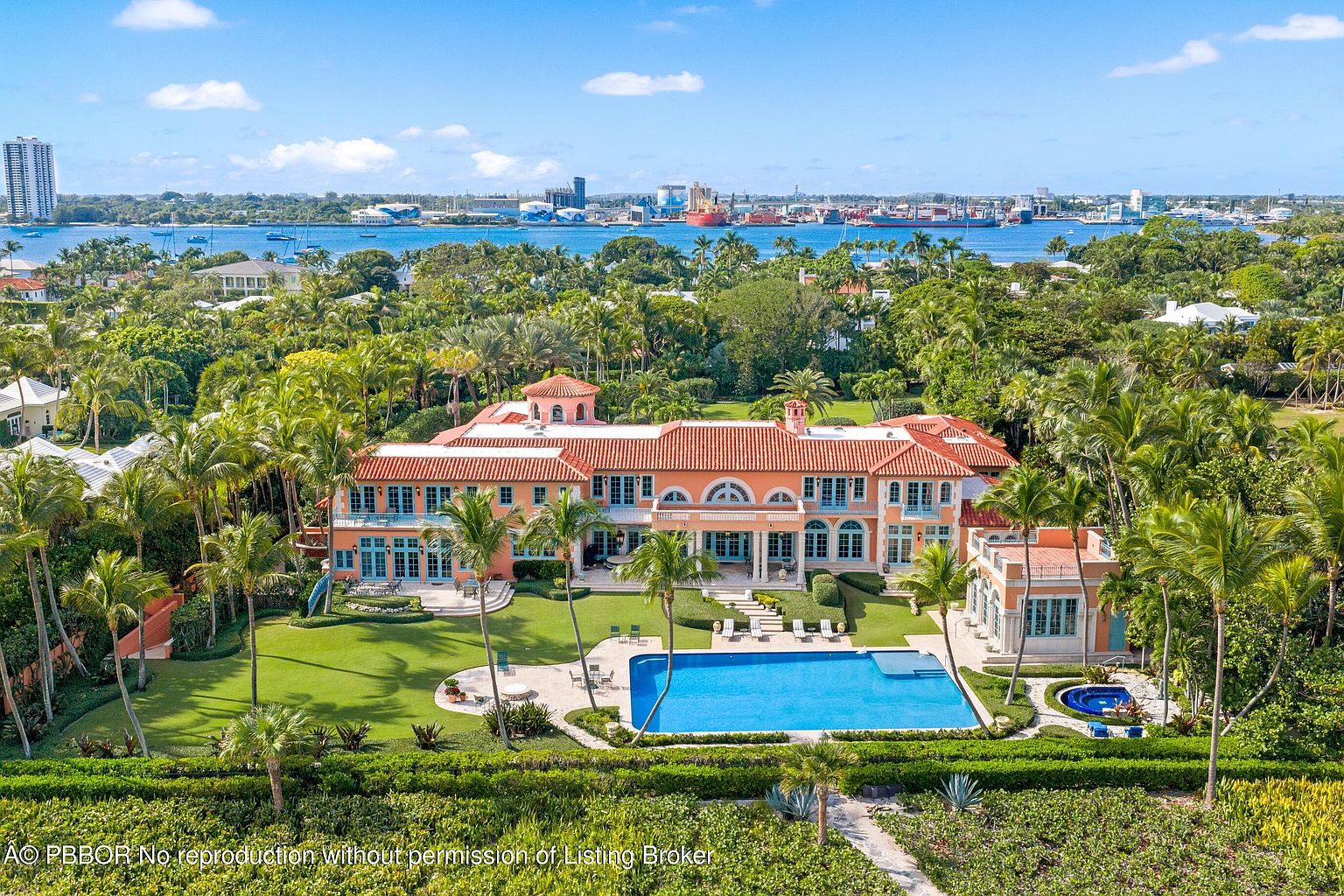 This aerial view showcases a grand estate with a large swimming pool, lush green lawns, and meticulously maintained gardens. The Mediterranean-style architecture features a terracotta roof, arched windows, and a symmetrical design. The property is surrounded by mature trees, offering privacy and a sense of seclusion, with a waterfront view in the background.