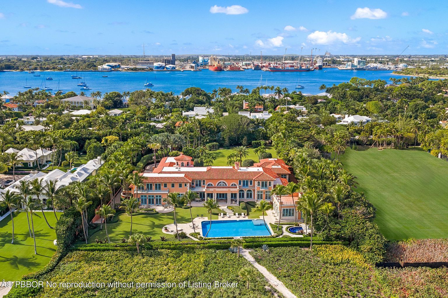 This aerial view showcases a grand estate with a large swimming pool, meticulously manicured lawns, and lush landscaping. The Mediterranean-style architecture features a terracotta roof, arched colonnades, and a symmetrical design. The property is situated near the waterfront, offering stunning views of the bay and distant cityscape.