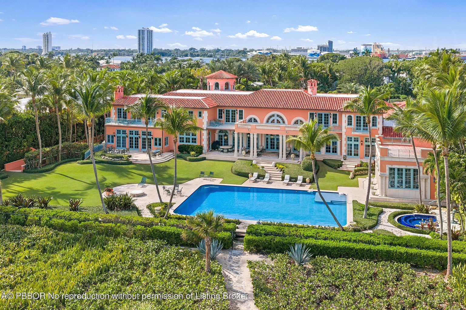 This aerial view showcases a grand estate with a terracotta roof, peach-colored facade, and multiple balconies. A large swimming pool is centrally located in the manicured lawn, surrounded by lush landscaping and palm trees. The property exudes luxury and privacy, with a glimpse of the city skyline in the distance.