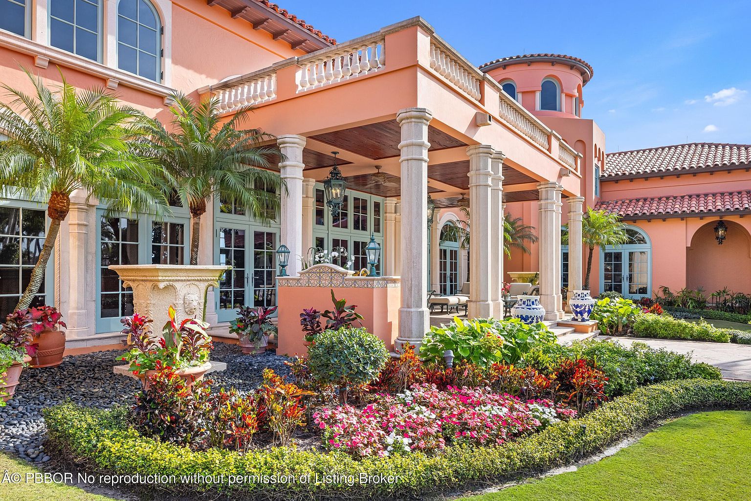 This image showcases a beautifully landscaped yard and garden area of a luxurious estate. The focal point is a vibrant flower bed with a variety of colorful blooms, complemented by lush greenery and manicured hedges. The architectural details of the building, including the columns and balcony, add to the overall elegance and curb appeal of the property.