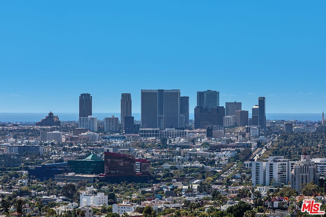 This aerial view showcases a sprawling cityscape under a clear blue sky, highlighting numerous high-rise buildings and a dense urban landscape. The perspective emphasizes the scale and density of the city, with residential areas and green spaces interspersed among the commercial structures. The image conveys a sense of urban living and the vastness of the metropolitan area.