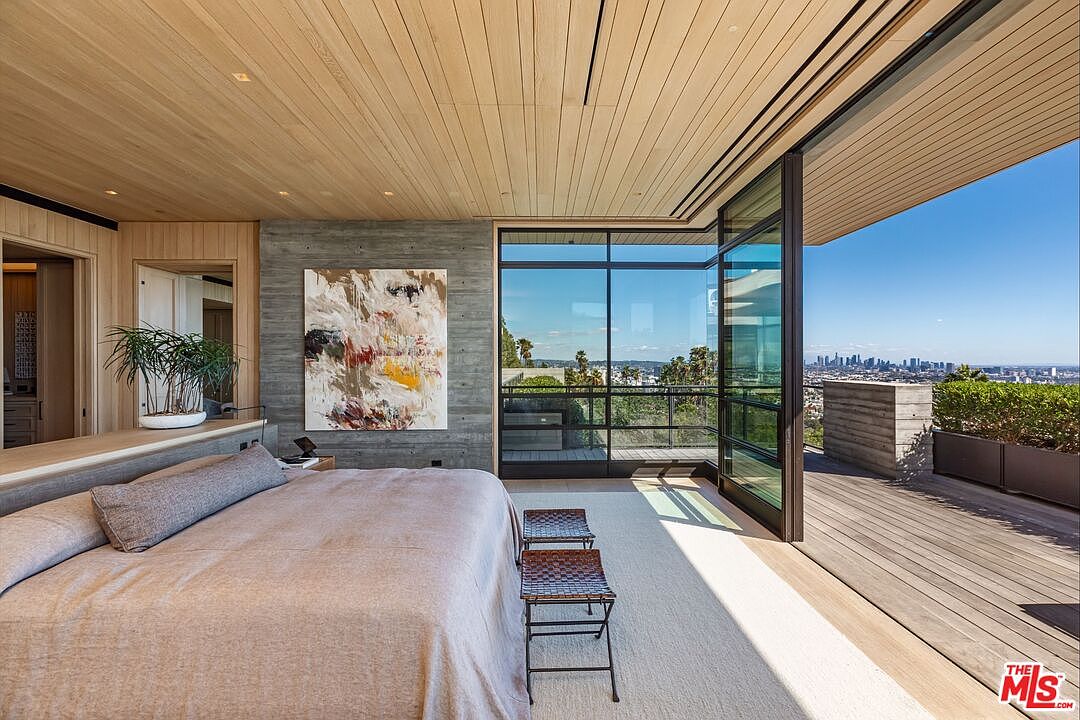 This is a luxurious primary bedroom featuring a king-size bed with neutral linens, an abstract art piece on a concrete wall, and floor-to-ceiling windows offering panoramic city views. The room has a modern design with wood paneling on the ceiling and walls, and it opens onto a spacious deck, creating a seamless indoor-outdoor living experience. The perspective is from the foot of the bed, looking towards the windows and the city skyline.