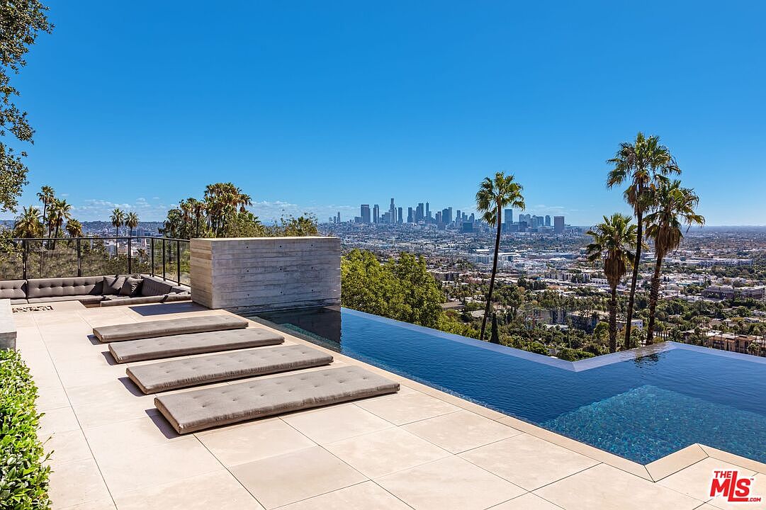 This image showcases a luxurious infinity pool with a stunning city view. The pool area features modern lounge pads and a sleek design, complemented by palm trees and clear blue skies. The overall impression is one of high-end relaxation and sophisticated outdoor living.