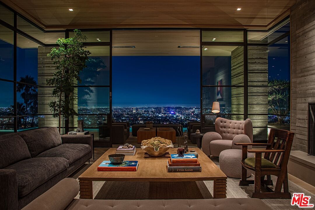 This is a luxurious living room featuring a modern design with floor-to-ceiling windows offering a stunning city view at night. The room is furnished with a comfortable gray sofa, a wooden coffee table with books, and a cozy armchair with an ottoman. The space is well-lit and exudes a sophisticated and inviting atmosphere.