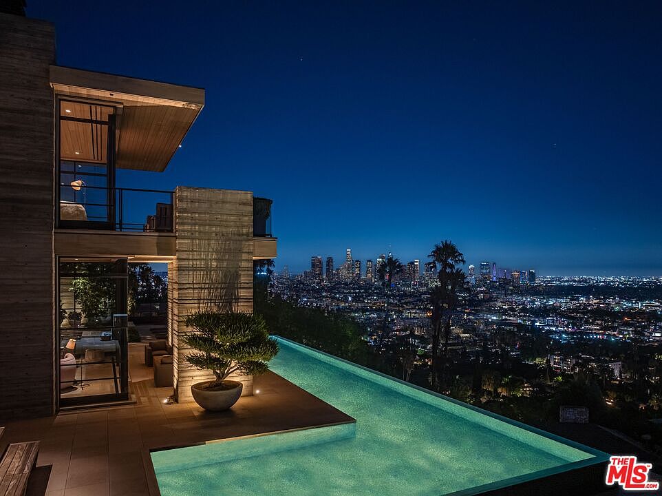 This image showcases a luxurious infinity pool at night, offering stunning city views. The pool is integrated into a modern architectural design, featuring clean lines and a sleek deck area with a small tree. The overall impression is one of high-end living and sophisticated outdoor space.