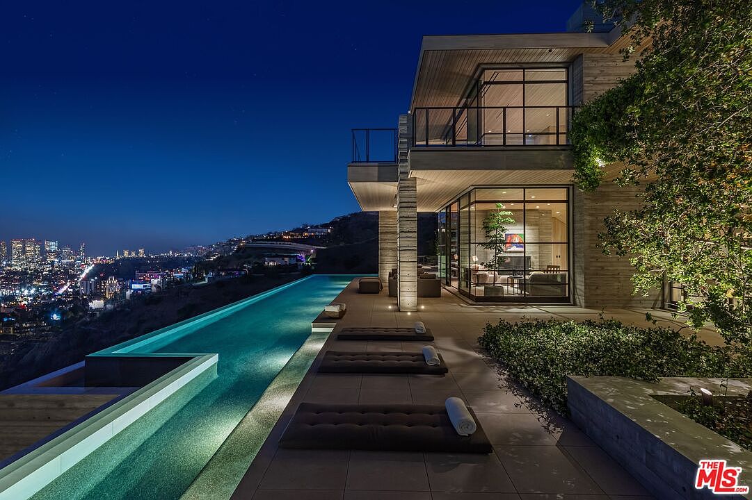 This image showcases a luxurious infinity pool at night, overlooking a stunning cityscape. The pool is surrounded by lounge chairs, and the modern architecture of the house features large windows that offer views of the interior. The overall impression is one of opulence and sophisticated design.