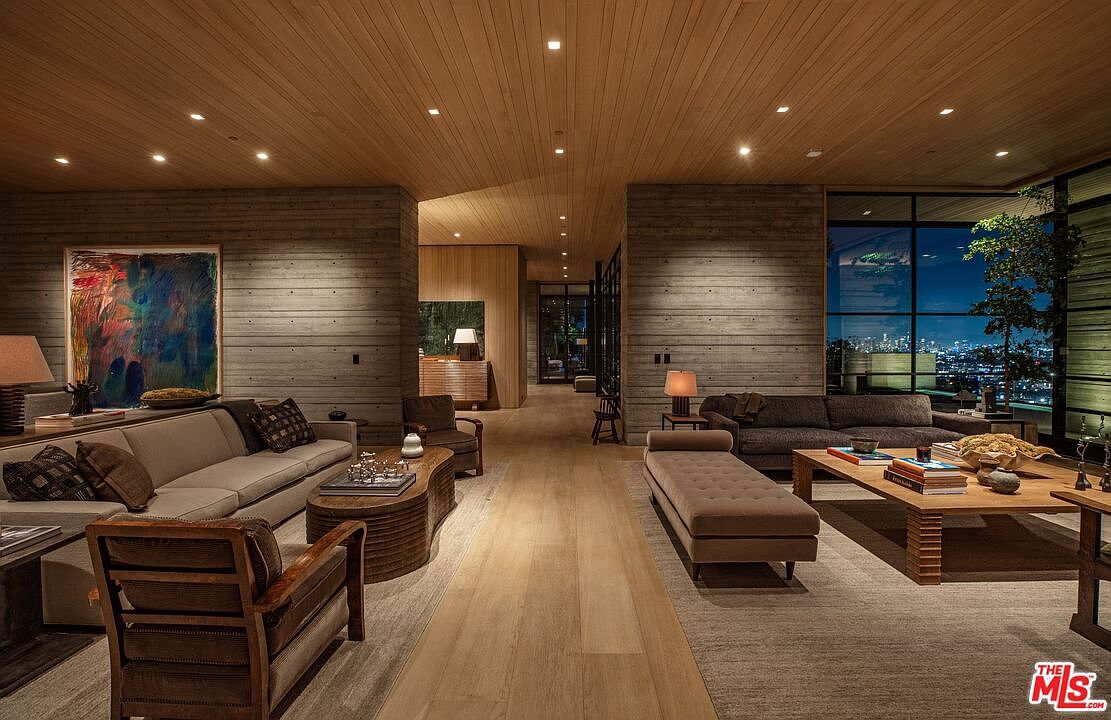 This is a spacious living room featuring modern design elements. The room is divided into two distinct seating areas, each with comfortable sofas and stylish coffee tables. Large windows offer a stunning city view, and the wood-paneled ceiling adds warmth to the space. The walls are concrete, and the floors are wood.