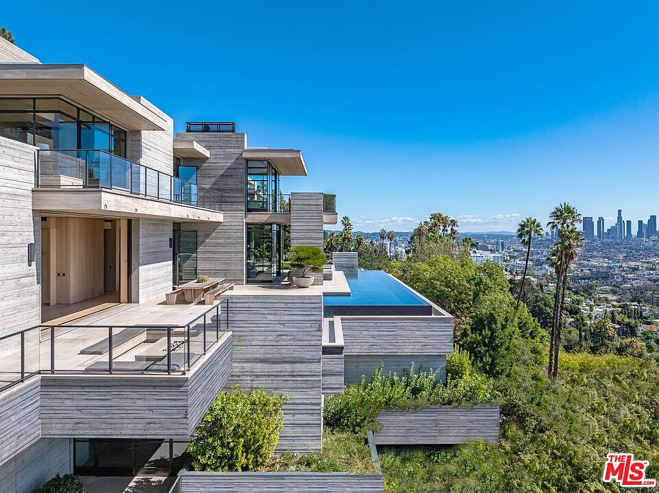 This image showcases the rear exterior of a modern, multi-level home constructed with concrete and glass. The design features multiple balconies, an infinity pool, and lush landscaping that blends seamlessly with the surrounding hillside. The view extends to the city skyline in the distance, emphasizing the property's prime location and luxurious appeal.