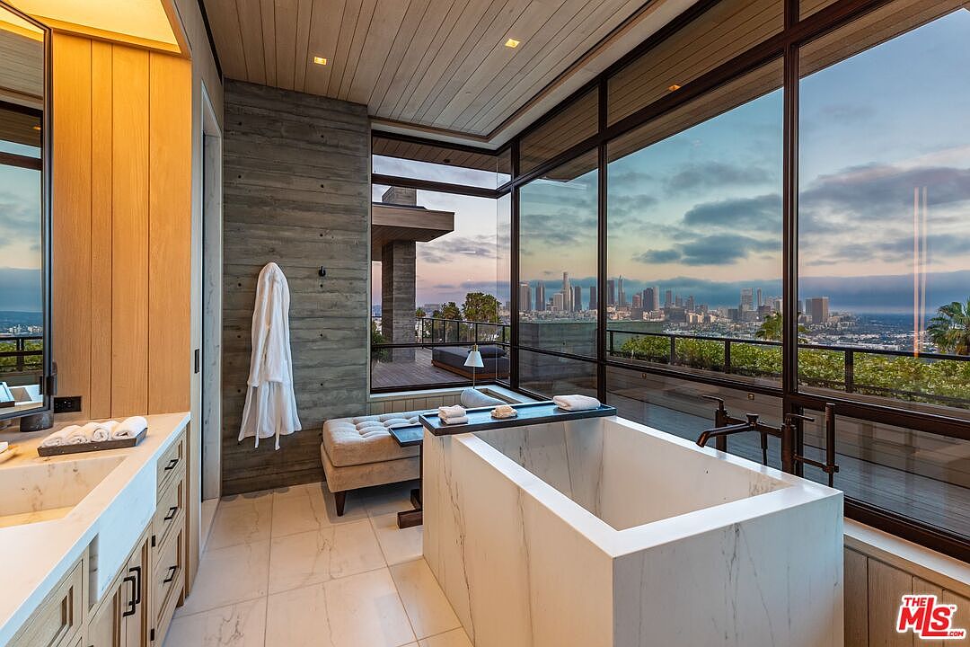 This is a luxurious primary bathroom featuring a modern, square-shaped marble bathtub positioned in front of large windows that offer a stunning city view. The bathroom includes a double vanity with marble countertops, wooden cabinetry, and a neutral color palette with wood and concrete accents. The space is designed for relaxation and showcases high-end finishes and fixtures.