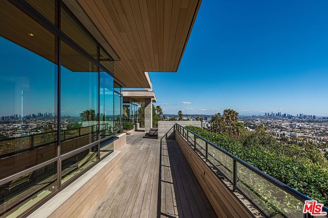 This image showcases a modern home's expansive deck or balcony, featuring sleek wooden flooring and a contemporary glass railing that offers unobstructed views of the cityscape. The architectural design emphasizes clean lines and open spaces, creating an inviting outdoor living area perfect for relaxation or entertaining. The large windows reflect the city skyline, blending the indoor and outdoor spaces seamlessly.