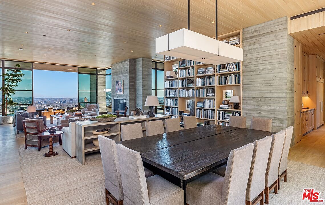 This interior shot showcases a luxurious dining area with a large, dark wood table surrounded by upholstered chairs. A striking rectangular light fixture hangs above the table, complementing the extensive built-in bookshelves filled with books. The room seamlessly transitions into a living area with floor-to-ceiling windows offering panoramic city views, creating an open and sophisticated living space.