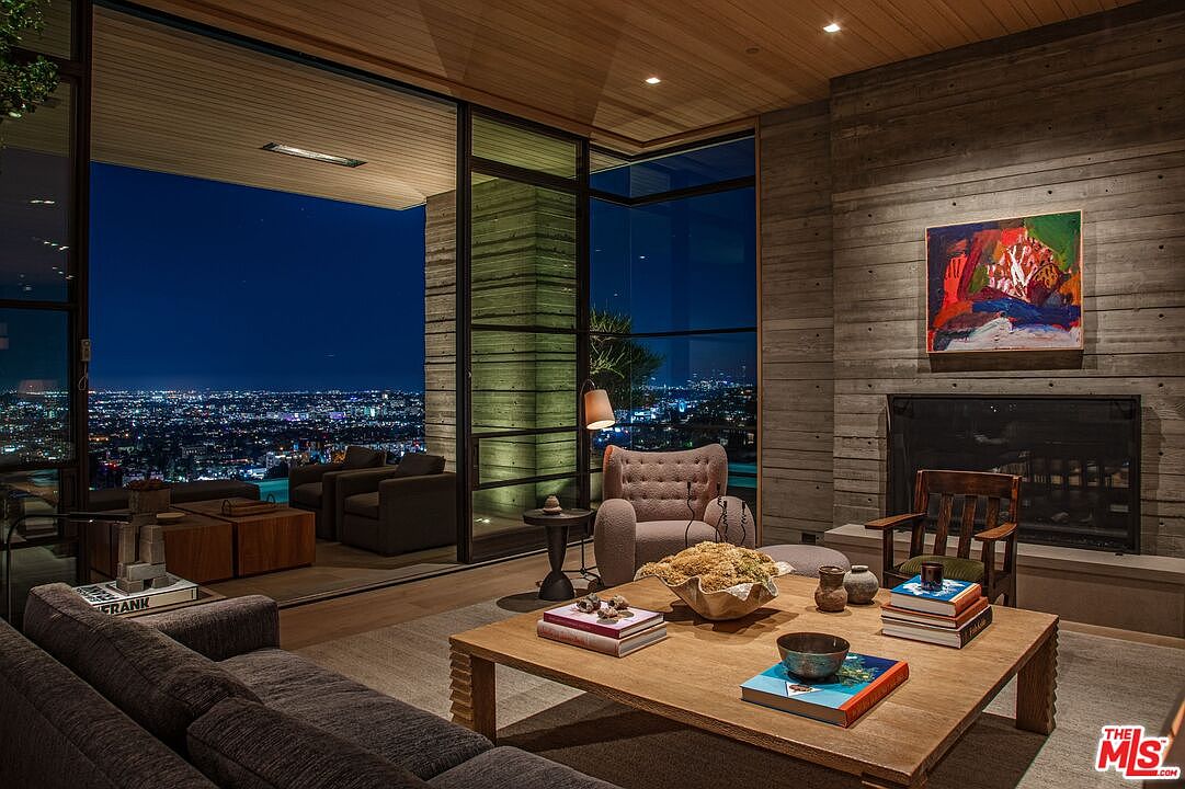 This is an interior shot of a luxurious living room featuring a modern design with concrete walls and wooden accents. Large windows offer a stunning nighttime city view, while the room is furnished with a comfortable sofa, armchairs, and a stylish coffee table adorned with books and decorative items. A fireplace adds warmth and ambiance to the space.