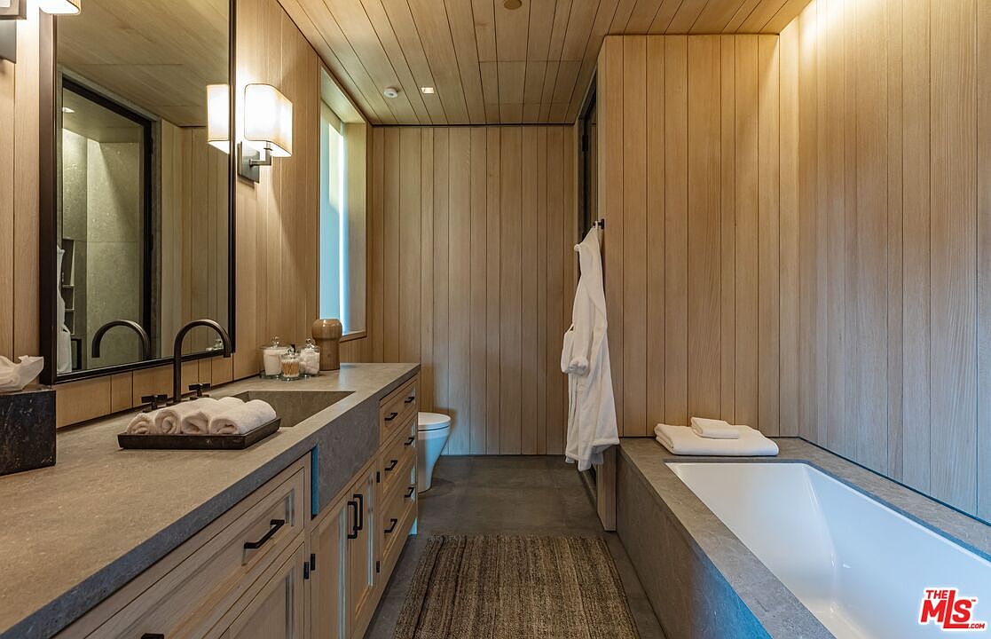 This is a primary bathroom featuring wood paneled walls and ceiling, creating a spa-like ambiance. The bathroom includes a long vanity with a concrete countertop and a rectangular sink, a large mirror, and a built-in bathtub with a concrete surround. A white robe hangs on the wall, adding to the luxurious feel of the space.