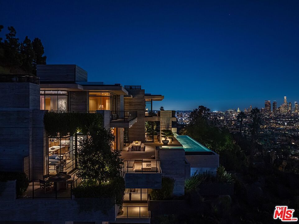 This image showcases the rear exterior of a modern luxury home at night, featuring multiple levels with balconies and a rooftop infinity pool. The architectural design emphasizes clean lines and a tiered structure, with warm interior lighting contrasting against the dark sky. The cityscape in the background adds to the property's appeal, highlighting its elevated location and stunning views.