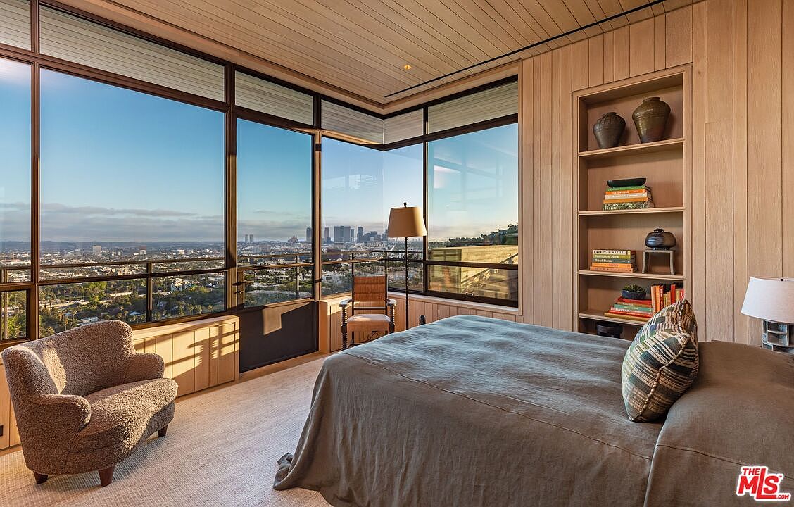 This is a primary bedroom featuring a large window with a city view, a cozy armchair, and a built-in bookshelf. The room has a modern and minimalist design with wood paneling and a neutral color palette. The bed is neatly made with a gray linen duvet and a decorative pillow.