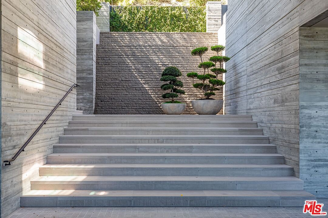 This image showcases a modern entryway with a grand staircase leading up to a textured stone wall adorned with meticulously shaped bonsai trees in minimalist concrete pots. The staircase is flanked by concrete walls with a horizontal wood grain pattern, adding a touch of contemporary elegance. The overall impression is one of sophisticated design and serene landscaping, creating an inviting and stylish entrance.