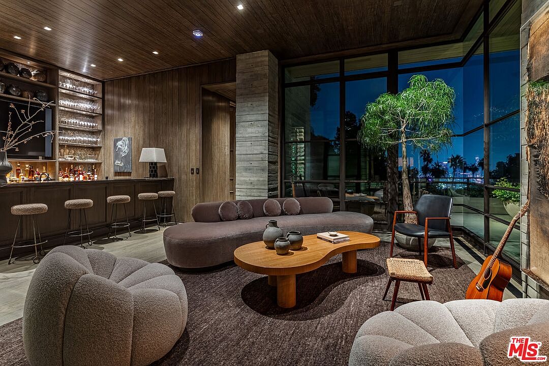 This is an interior shot of a luxurious living room featuring a bar area, a modern curved sofa, and unique rounded armchairs. The room is illuminated by recessed lighting and natural light from large windows, offering a view of the city skyline at night. A guitar leans against the wall, adding a touch of personality to the space.
