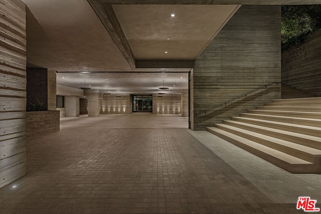 This image showcases a grand entryway featuring a modern architectural design with concrete and wood-like textures. The space is open and spacious, with a tiled floor leading into an interior area illuminated by recessed lighting and pendant fixtures. A staircase with a metal railing ascends to the right, adding to the impressive scale of the entrance.
