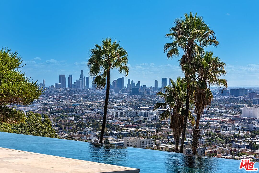 This image showcases an infinity pool with a stunning panoramic view of a cityscape, likely Los Angeles, featuring palm trees in the foreground. The pool's edge seamlessly blends with the horizon, creating a luxurious and serene atmosphere. The clear blue sky enhances the sense of openness and exclusivity, making it an attractive feature for a high-end property.