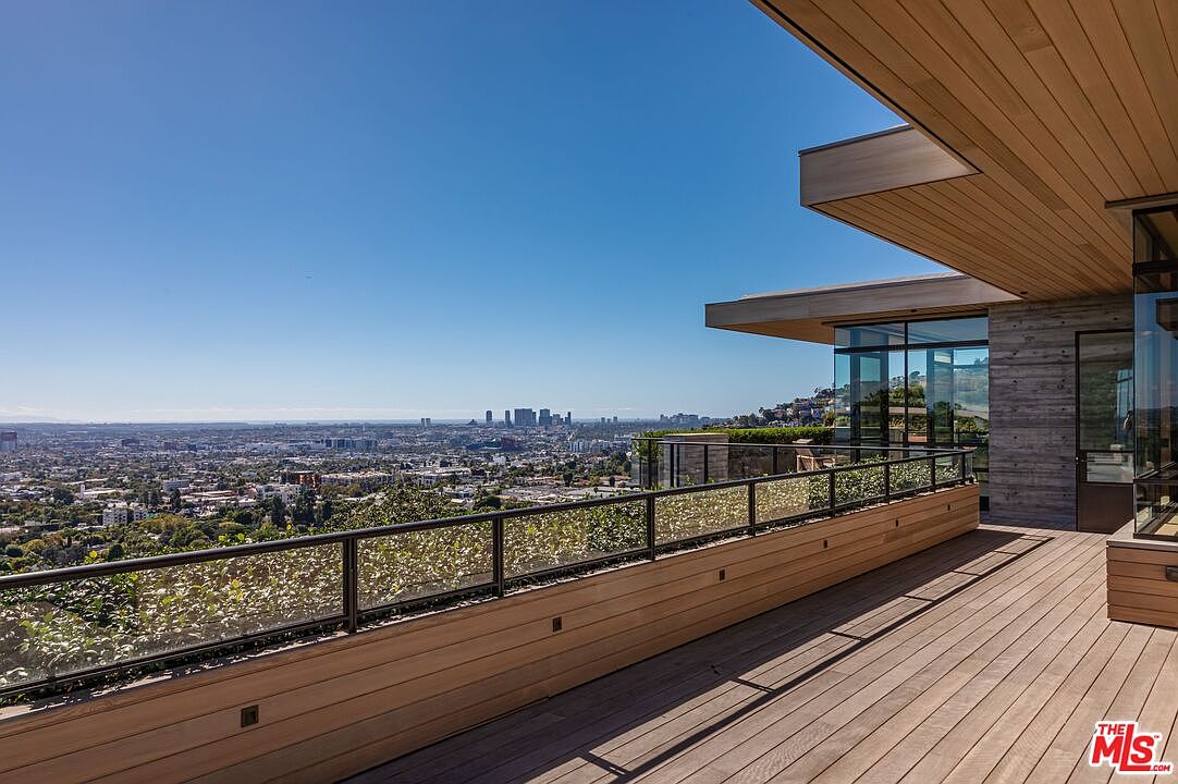 This image showcases a luxurious patio or deck area with a stunning panoramic view of a cityscape. The deck features wooden flooring and a modern glass railing with greenery, offering both privacy and an unobstructed view. The architectural design of the building is contemporary, with clean lines and a combination of wood and concrete elements.