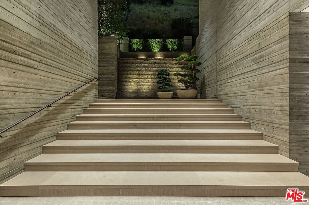 This image showcases an impressive entryway with wide, light-colored stone steps leading up to a landscaped area. The walls on either side are constructed of horizontally layered concrete, adding a modern and textured feel. Two potted bonsai trees flank the top of the stairs, illuminated by subtle lighting, creating an inviting and sophisticated entrance.