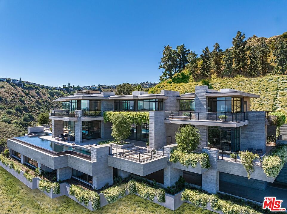 This aerial view showcases a modern, multi-level luxury home nestled into a hillside. The architectural design features clean lines, concrete elements, and expansive glass windows, complemented by lush greenery and a rooftop pool. The property offers stunning views and a seamless integration with the natural landscape.