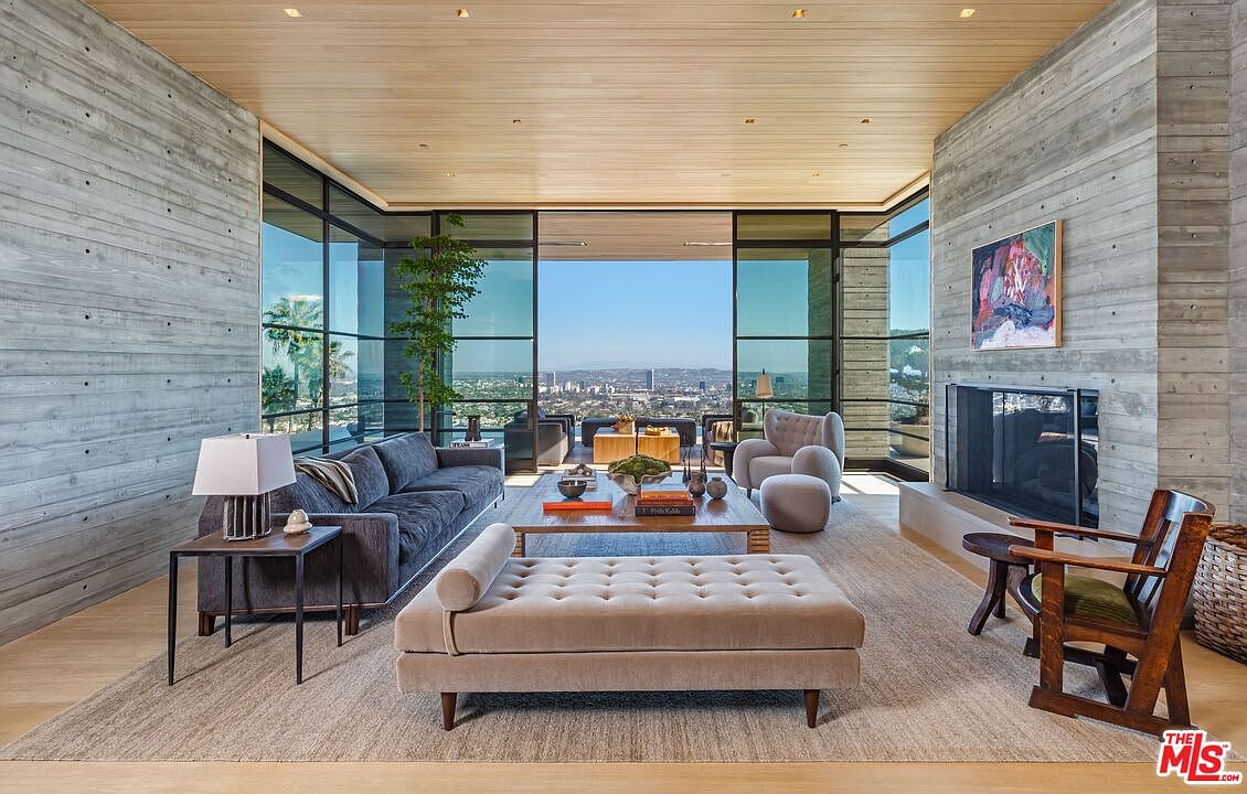 This is a spacious and modern living room featuring concrete walls, a wood plank ceiling, and floor-to-ceiling windows offering a panoramic city view. The room is furnished with a gray sofa, a beige chaise lounge, a wooden coffee table, and two wooden chairs near the fireplace, creating a comfortable and stylish living space. The open layout and natural light enhance the room's appeal.