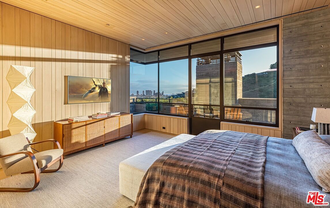 This primary bedroom features wood paneled walls, a large window with city views, and a modern aesthetic. A media console sits beneath a wall-mounted television, and a comfortable bed with a striped blanket anchors the space. The room is bathed in natural light, creating a serene and inviting atmosphere.