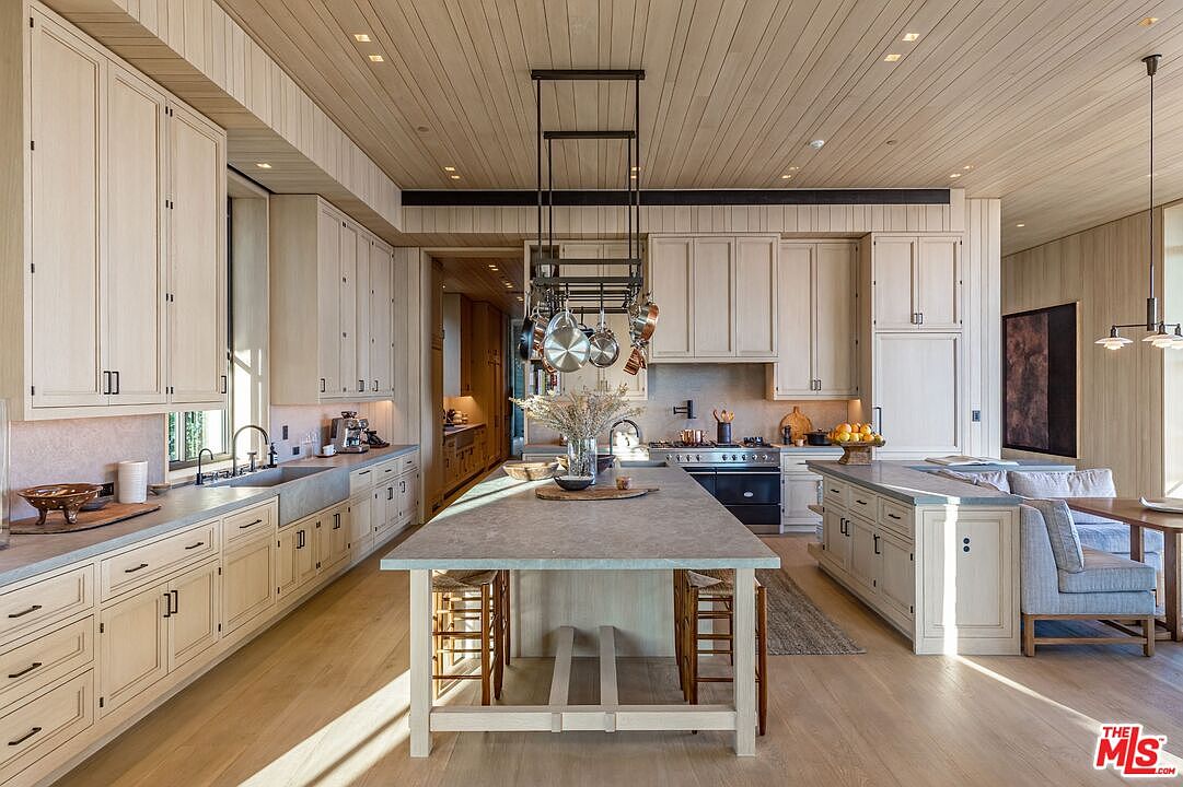 This is a spacious and well-lit kitchen featuring light-colored wooden cabinetry and a large central island with seating. The kitchen is equipped with modern appliances and a hanging pot rack above the island. The overall design is elegant and functional, creating a warm and inviting atmosphere.